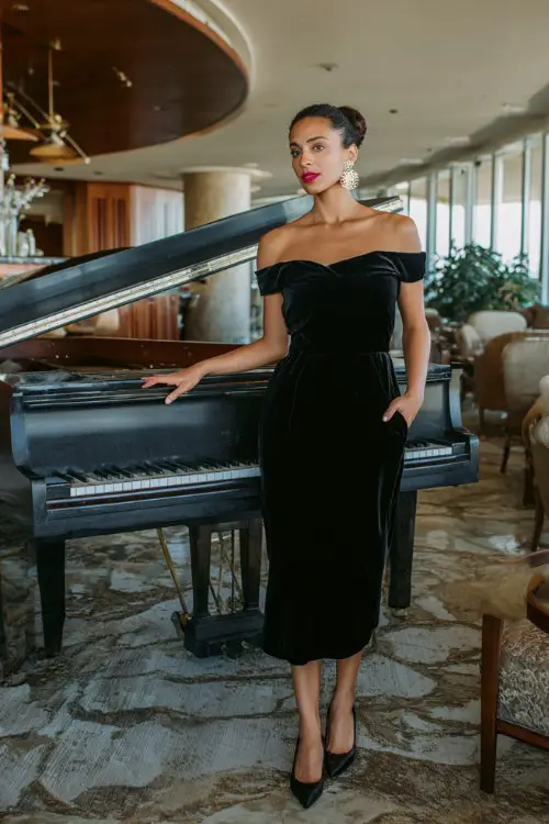 A woman wears a black velvet off-shoulder dress with diamond studs, classic pumps, and a low bun