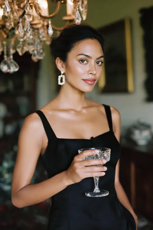 A woman wears an old money party outfit with black silk midi dress with a square neckline, pearl earrings