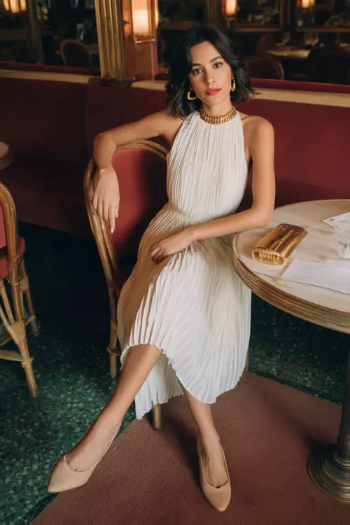 A woman wears an old money style date night outfit with a white pleated midi dress with a bateau neckline, paired with beige pumps, gold hoops, and a small clutch
