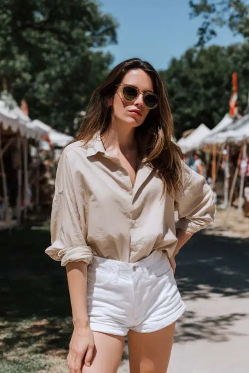 woman wearing oversized beige button up shirt with rolled sleeves, high waisted white denim shorts, white sneakers and aviator sunglasses