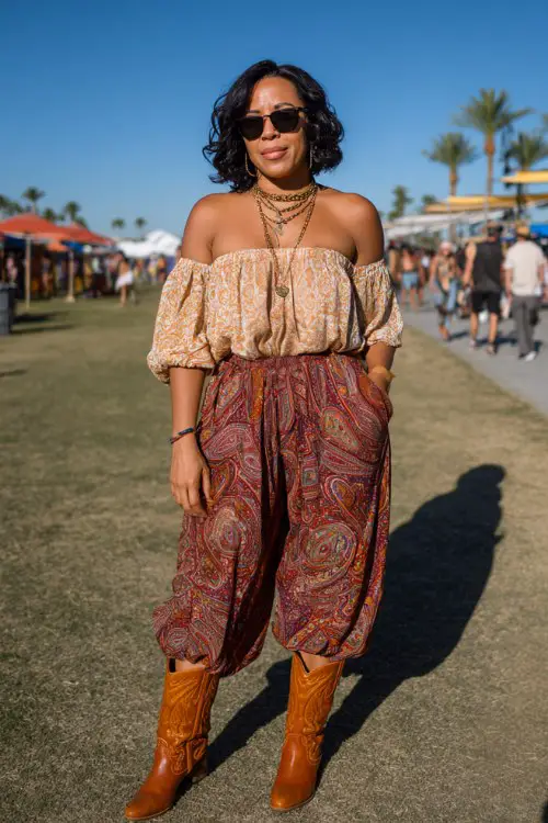 A curvy woman wears caramel cowboy boots with wide-leg paisley pants and a breezy off-shoulder blouse, accessorized with layered necklaces and oversized sunglasses