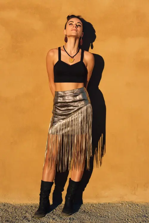 A woman wearing a fitted black crop top, metallic fringe skirt, statement earrings, and black boots, plain sunlit ground background