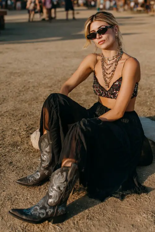 A woman wears metallic cowboy boots with a beaded crop top and flowing chiffon maxi skirt, styled with layered chains and oversized sunglasses