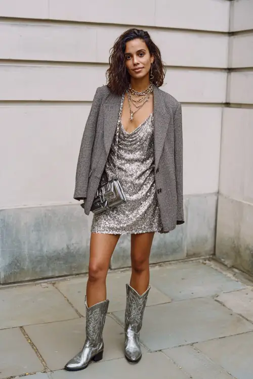 A woman wears silver cowboy boots with a shimmering sequin slip dress and oversized blazer, styled with layered chains and a structured mini bag