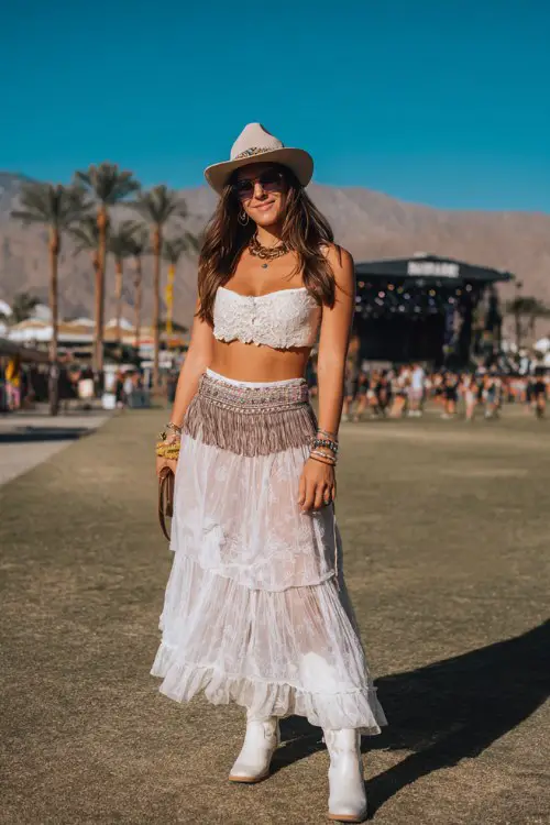 A woman wears white cowboy boots with a tiered chiffon maxi skirt and lace-trim crop blouse, styled with layered bracelets and a fringe belt bag