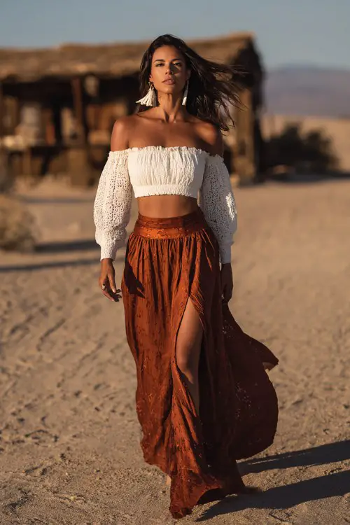 a woman walking in a desert, wearing a white crochet off-shoulder crop top and a high-waisted rust brown boho maxi skirt with a thigh slit