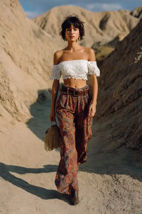 stylish model walking through a sandy trail, wearing a white crochet off-shoulder crop top and high waisted paisley wide-leg pants, leather belt, fringe handbag