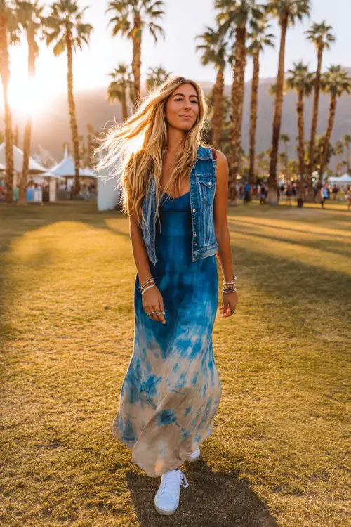 tall slim woman walking through Coachella festival grounds in the desert, wearing a blue and beige tie-dye maxi dress with a sleeveless denim vest, white sneakers