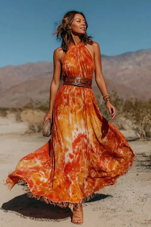 woman wearing a flowing tie-dye maxi dress with halter neckline and fringe hem, brown leather belt at the waist, sandals