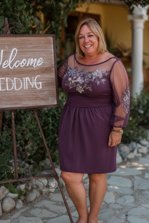 A plus-size woman over 50 wearing a plum-colored knee-length dress with sheer sleeves and embroidered floral accents, posing by a wedding welcome sign