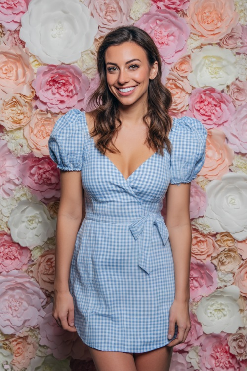A woman in a blue and white gingham short dress with puff sleeves and tie waist, standing beside a DIY wedding photo booth with paper florals 