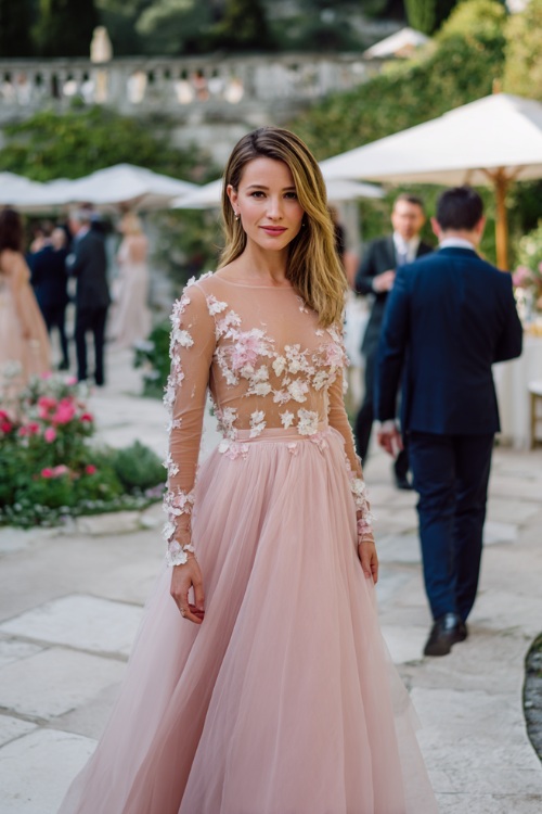 A woman in a blush pink tulle gown with sheer long sleeves, floral appliqué details, and a sweeping skirt, walking slowly through a garden reception