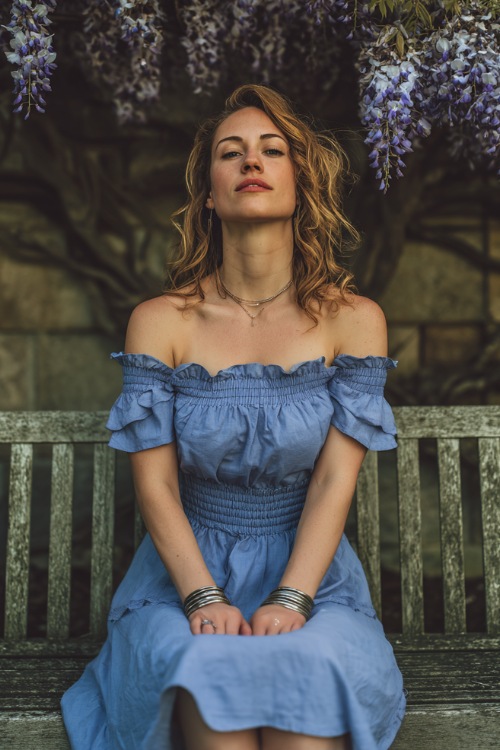 A woman in a dusty blue off-the-shoulder midi dress with ruffle trim, silver bangles, and soft waves, seated on a bench under blooming wisteria