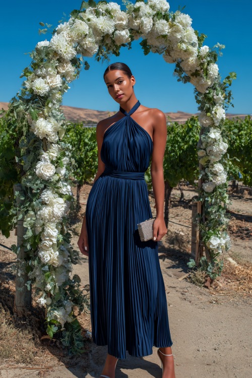 A woman in a navy blue halter-neck pleated maxi dress with silver heels and a matching clutch, standing beside a white floral wedding arch in a sunny vineyard 