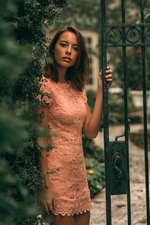 A woman in a peach knee-length lace dress with scalloped edges and short sleeves, standing beside a wrought-iron garden gate covered in greenery