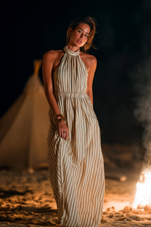 A woman in a striped beige and white cotton dress with a high neckline and relaxed shape, standing barefoot near a beach wedding bonfire area 