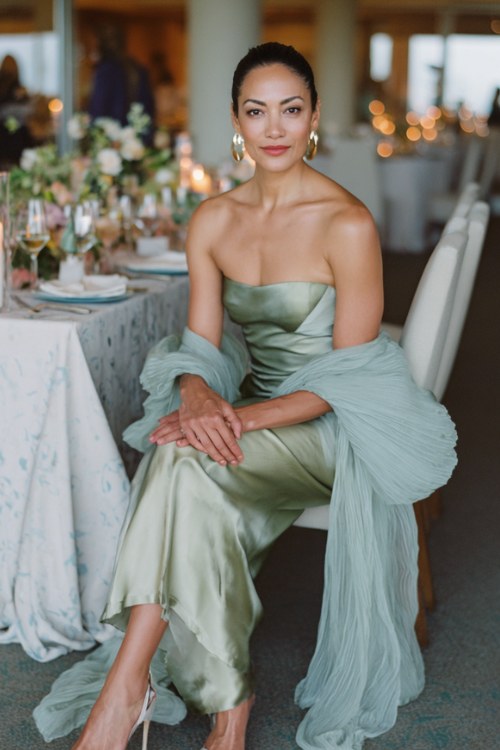 A woman over 40 wears a sage green strapless formal gown with a dramatic pleated cape of crinkled silk organza fanning from the upper back like wings, the front sleek and unadorned