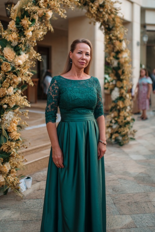A woman over 50 in a dark green floor-length formal gown with lace three-quarter sleeves and a fitted waist, standing beside a grand wedding arch under golden light 