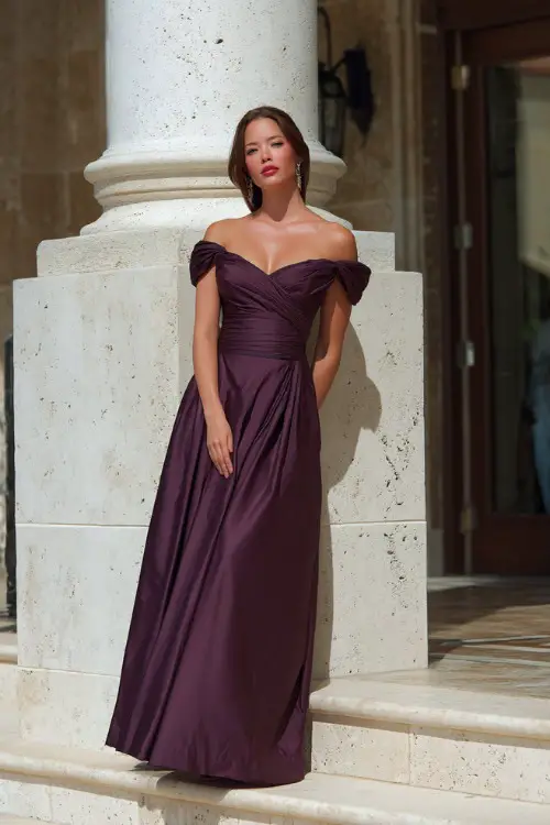 A woman over 50 in a deep plum evening gown with off-the-shoulder sleeves and a floor-length skirt, standing on the steps of a formal outdoor wedding venue 