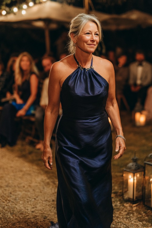 A woman over 50 in a midnight blue evening gown with a high neckline, fitted bodice, and gentle flare at the hem, walking through a candlelit outdoor wedding aisle 