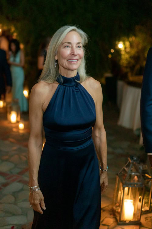 A woman over 50 in a midnight blue evening gown with a high neckline, fitted bodice, and gentle flare at the hem, walking through a candlelit outdoor wedding aisle