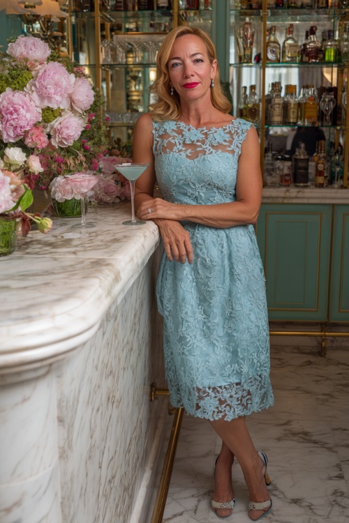 A woman over 50 wearing a sky-blue tea-length dress with floral lace overlay and scalloped hem, posing beside a wedding cocktail bar decorated with peonies (2)