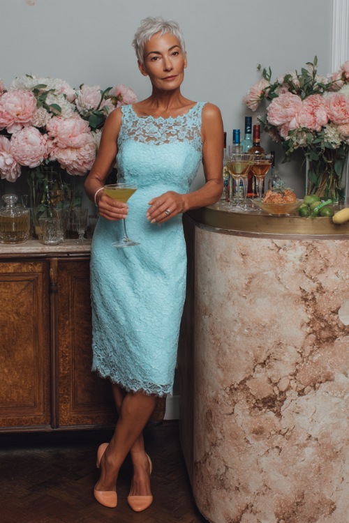 A woman over 50 wearing a sky-blue tea-length dress with floral lace overlay and scalloped hem, posing beside a wedding cocktail bar decorated with peonies 