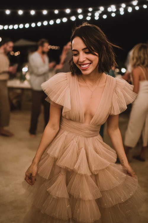 A woman wearing a tulle layered blush dress with a flowy silhouette and short sleeves, dancing beside a wedding reception under string lights 