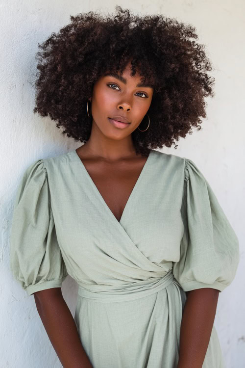 Black woman with voluminous curls wearing a soft sage green wrap dress with puff sleeves and minimalist jewelry, standing confidently in front of a clean white studio wall