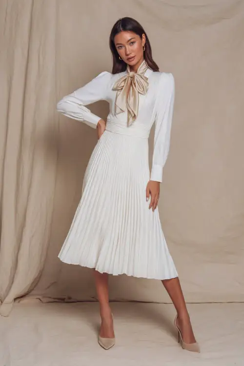 Elegant lady in a white pleated midi dress with a silk scarf tied around her neck, posing confidently in a studio with a soft matte cream backdrop