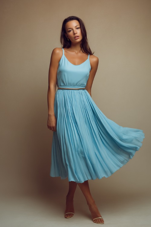 Elegant woman wearing a light blue sleeveless midi dress with pleated skirt and a thin belt, posing gracefully against a soft taupe studio backdrop 