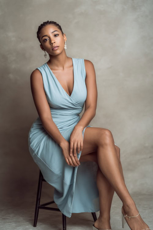 Graceful Black woman in a light blue sleeveless midi wrap dress with delicate drop earrings and classic low heels, posing elegantly in a minimalist studio with soft light stone backdrop 