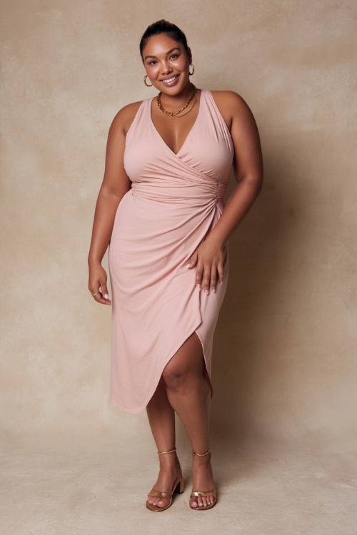 Plus-size woman wearing a soft pastel pink sleeveless wrap midi dress with delicate gold jewelry and block-heel sandals, posing gracefully against a soft beige studio backdrop