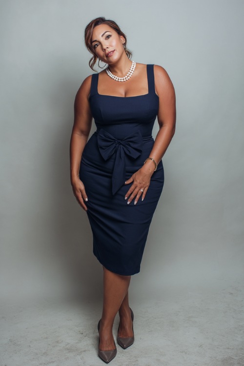 Sophisticated plus-size woman in a navy sleeveless midi dress with a square neckline, delicate pearl necklace, and classic heels, posing in front of a minimalist pale gray studio backdrop