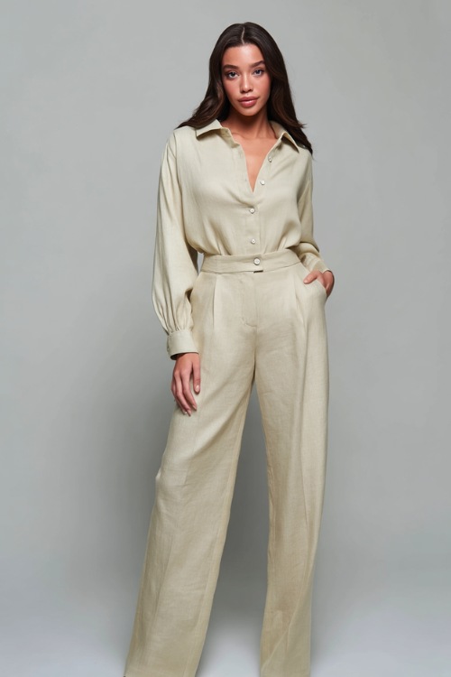 Woman wearing a high-waisted linen wide-leg trouser set in pale sage with a matching long-sleeve blouse, standing in a high-end studio with a minimalist soft gray background