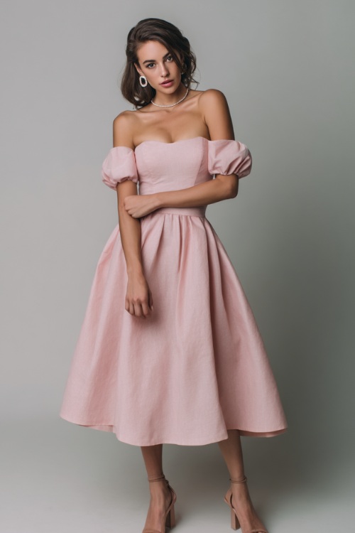 Woman wearing a soft pastel pink off-the-shoulder tea-length dress with subtle jewelry and block heels, posing elegantly in a minimalist fashion studio with a pale gray backdrop