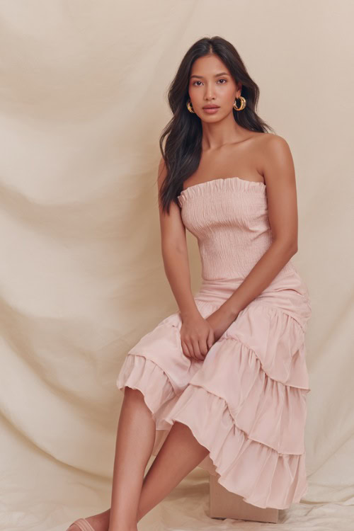 Woman wearing a soft pastel pink ruffled midi dress with delicate gold hoop earrings and strappy sandals, posing gracefully against a soft cream studio backdrop 