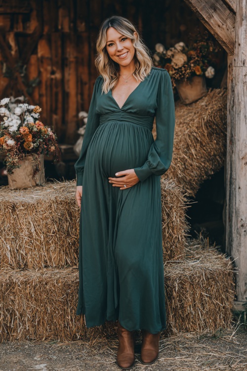 A woman wearing a flowy maternity-style forest green maxi dress with long sleeves and empire waist, styled with ankle boots, posing near a barn with hay bales and fall florals