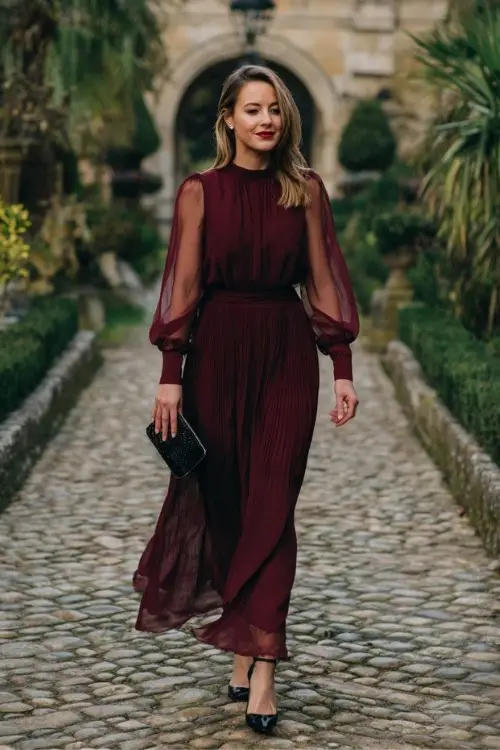 A woman wears a burgundy chiffon maxi dress with sheer bishop sleeves and a pleated skirt, paired with black heels and a satin clutch