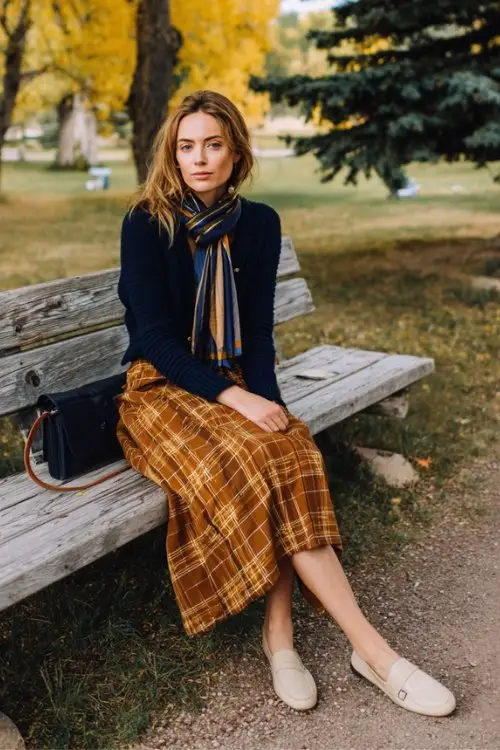 A woman wears a plaid midi skirt in warm tones with a navy cashmere cardigan, styled with loafers and a silk scarf