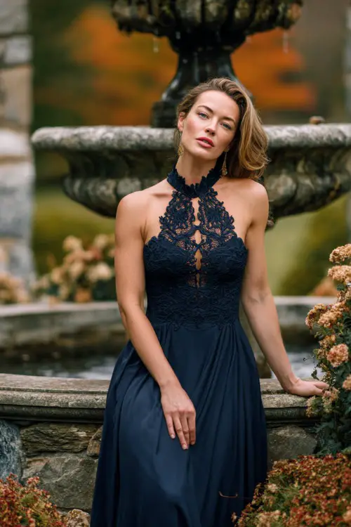 Elegant mature woman in a navy high-neck gown with lace bodice and chiffon skirt, posing beside a romantic stone fountain surrounded by fall florals