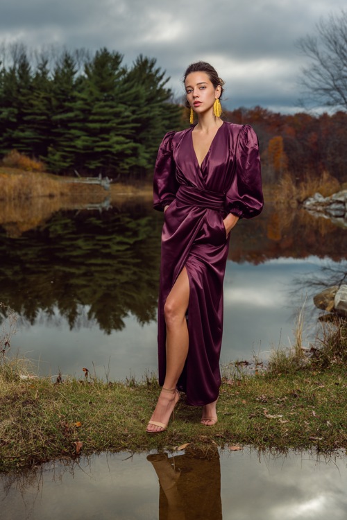 Elegant woman in a plum satin dress with bishop sleeves and a high slit, gold earrings and heeled sandals, standing by a reflective lake surrounded by late-autumn trees 