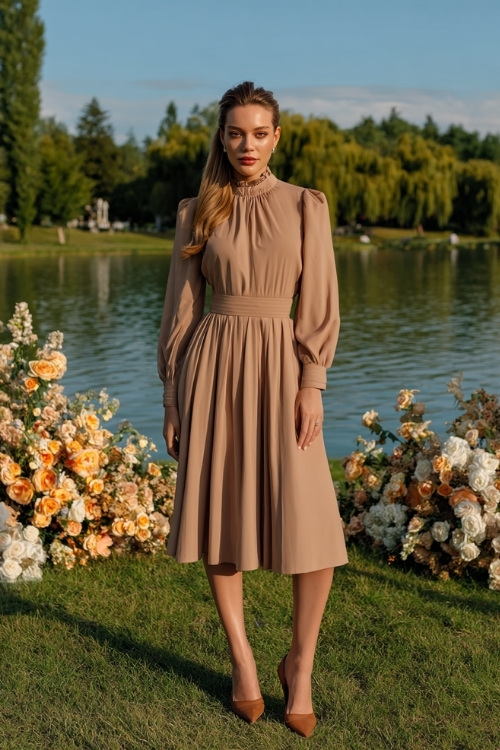 Elegant woman in a taupe high-neck midi dress with bishop sleeves and pleated skirt, paired with suede heels, posing by a lake surrounded by amber trees and floral aisle décor 