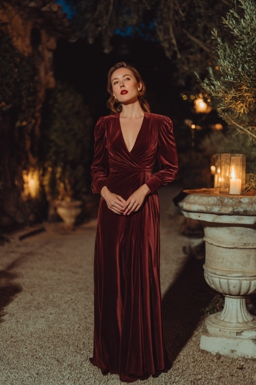 Elegant woman wearing a burgundy velvet maxi dress with long fitted sleeves and a modest v-neckline, standing at a candlelit fall wedding in a historic estate garden 