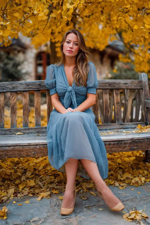Full body of a mature woman in a dusty blue midi dress with soft chiffon overlay and bow-tied neckline, surrounded by crisp golden leaves and wooden benches