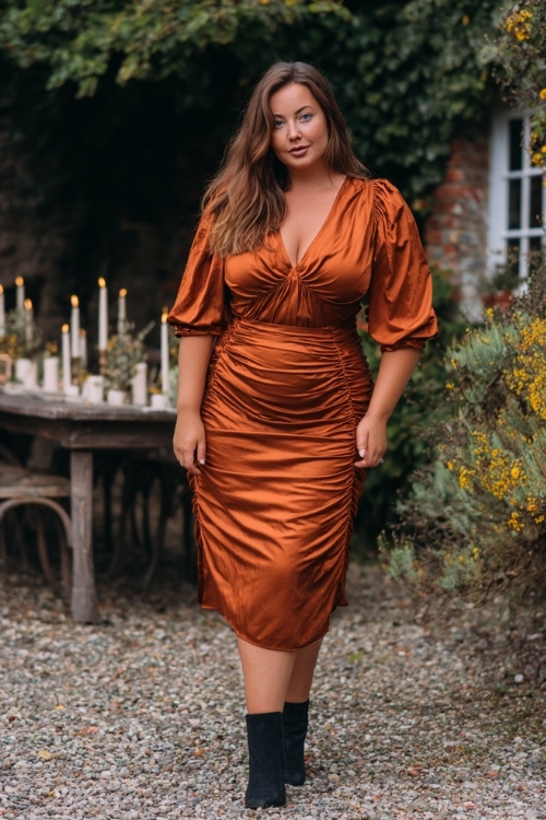 Full body of a plus-size woman in a burnt copper midi dress with puff sleeves and ruched detailing, styled with black ankle boots, standing in a small garden wedding with candles and foliage 