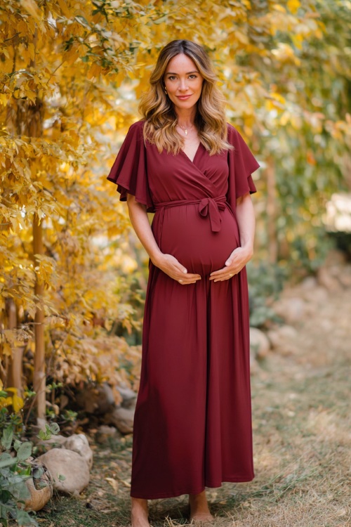 Full body of a woman in a bump-friendly burgundy wrap midi dress with flutter sleeves and a soft tie waist, standing in a golden-leafed garden at a rustic October wedding 