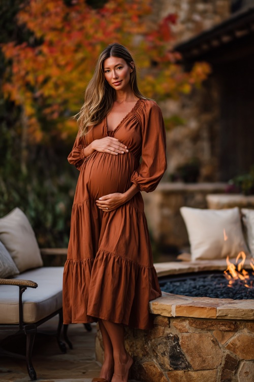 Full body of a woman in a cinnamon-colored maternity-friendly midi dress with tiered skirt and smocked bodice, posing near a firepit lounge with autumn foliage 