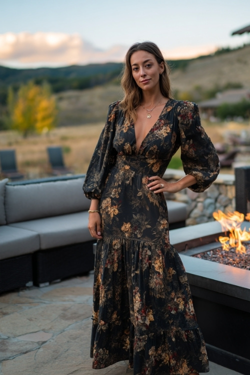 Full body of a woman in a dark floral midi dress with balloon sleeves and tiered skirt, standing near a firepit lounge at an outdoor fall wedding reception 