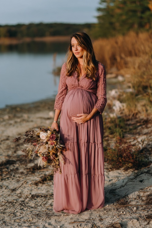 Full body of a woman in a dusty rose chiffon maternity-style gown with tiered skirt and lace bodice, standing near a lakeside October wedding with dried florals